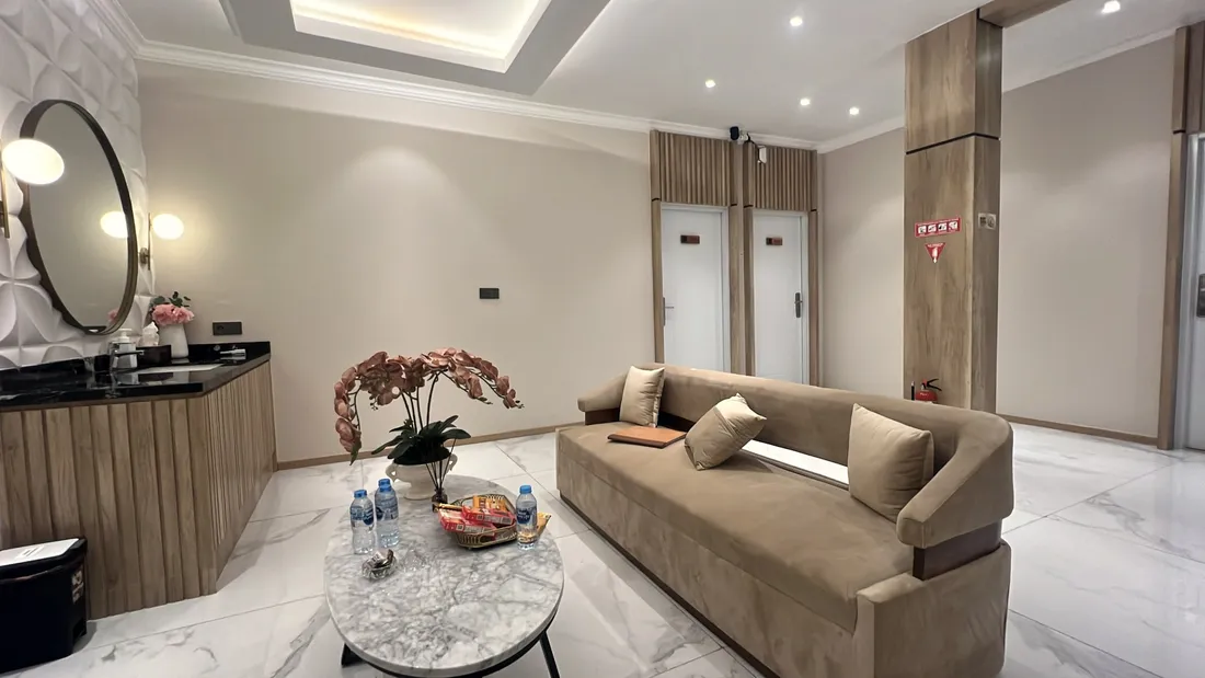 Ruang tunggu dengan sofa beludru krem, meja marmer bundar, anggrek, dan snack