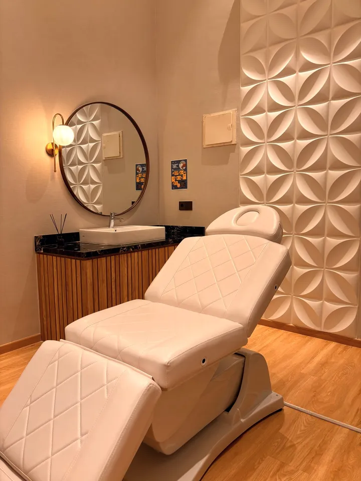 Ruang perawatan privat dengan bed treatment putih, cermin bundar berbingkai, dan dinding panel bunga 3D